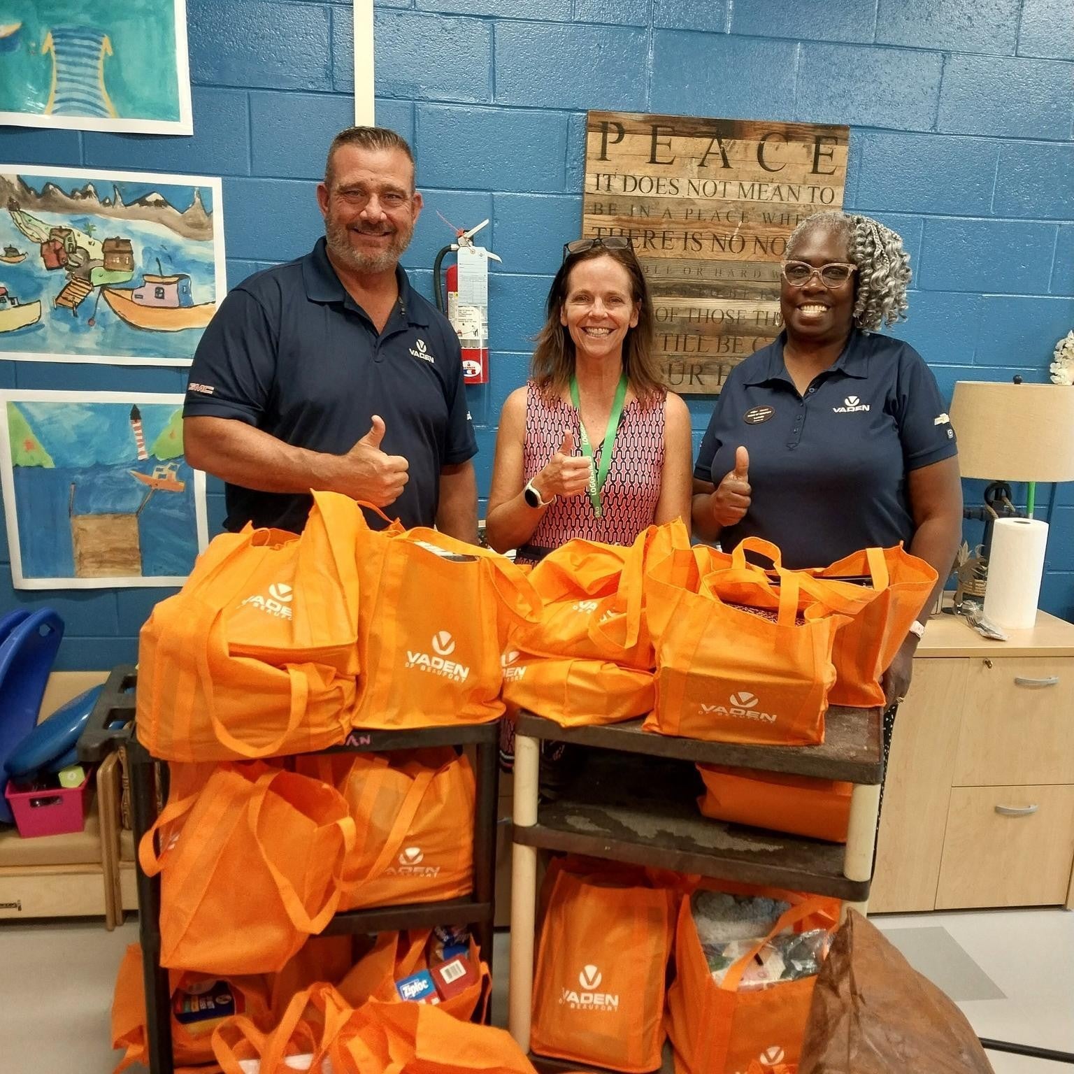 Three people giving thumbs ups while standing behind a two tier cart of bright orange bags with items in them.